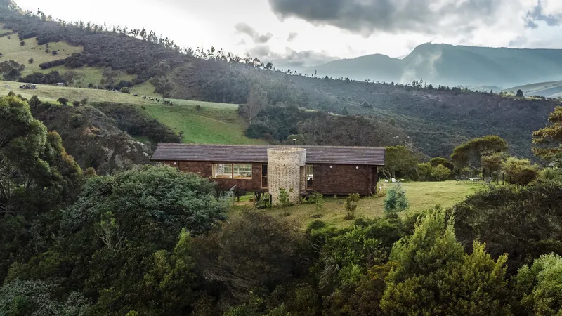 Horizontal timber house with brick tower nestled in sloping pasture beneath mountain ridges