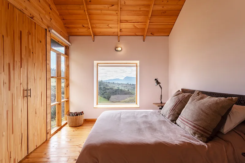 Bedroom with exposed timber ceiling, pine flooring and square window framing the distant hillside