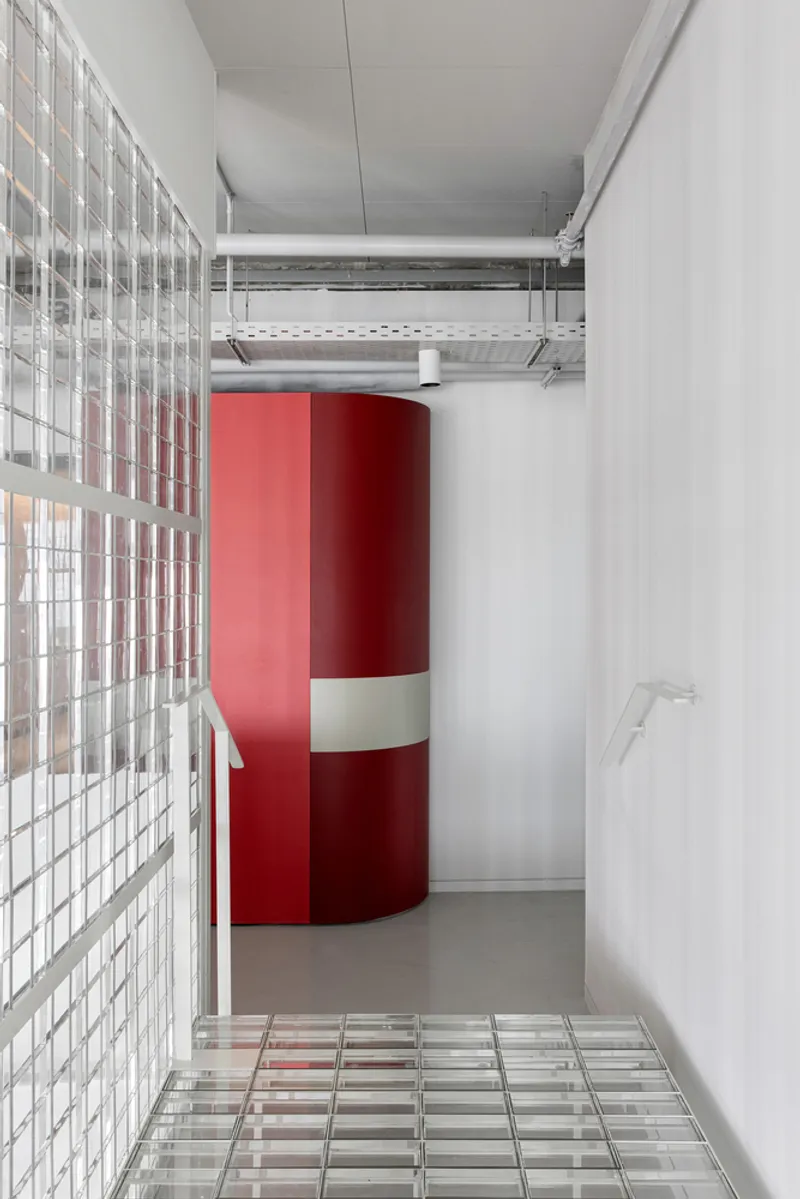 Corridor with glass block wall, red cylindrical column, and exposed ceiling ducts above glass tile flooring