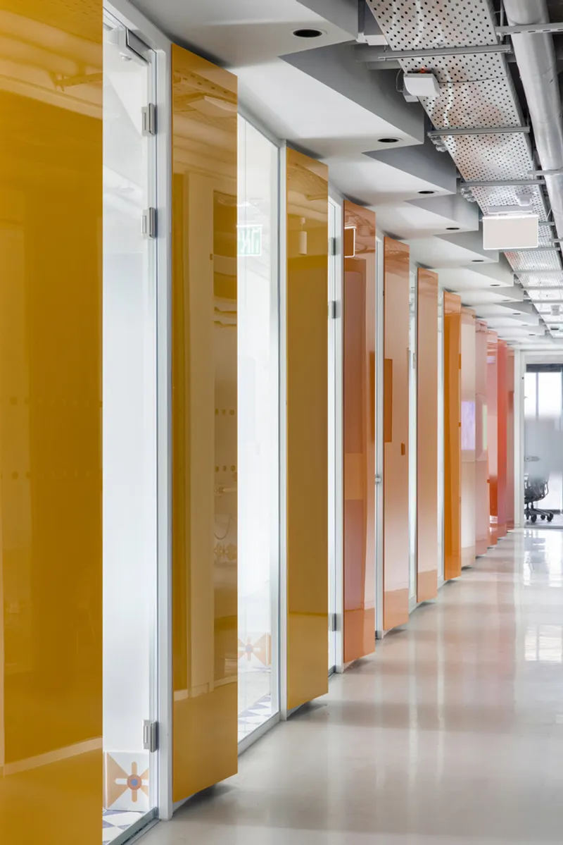 Interior corridor lined with glass-doored rooms featuring gradient color panels from yellow to coral and an exposed ceiling