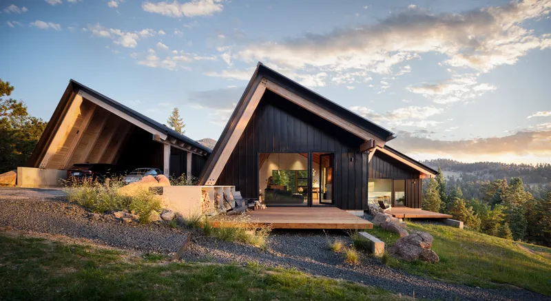 Two gabled volumes with dark metal cladding and timber-lined soffits on a gravel hillside at sunset