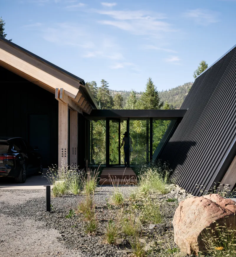 Courtyard view with black corrugated volumes framing a glazed corridor and gravel landscaping with native grasses