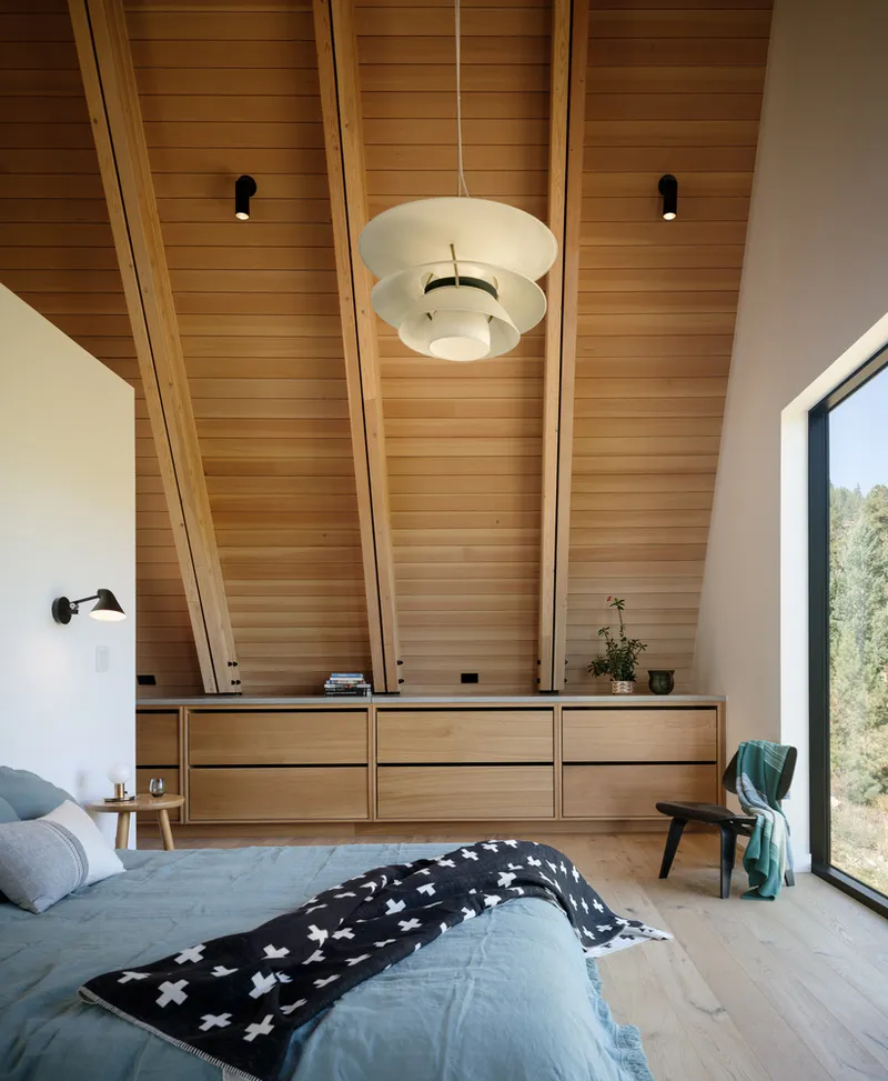 Bedroom with layered wood ceiling planes, pendant light, built-in storage wall, and floor-to-ceiling window overlooking trees
