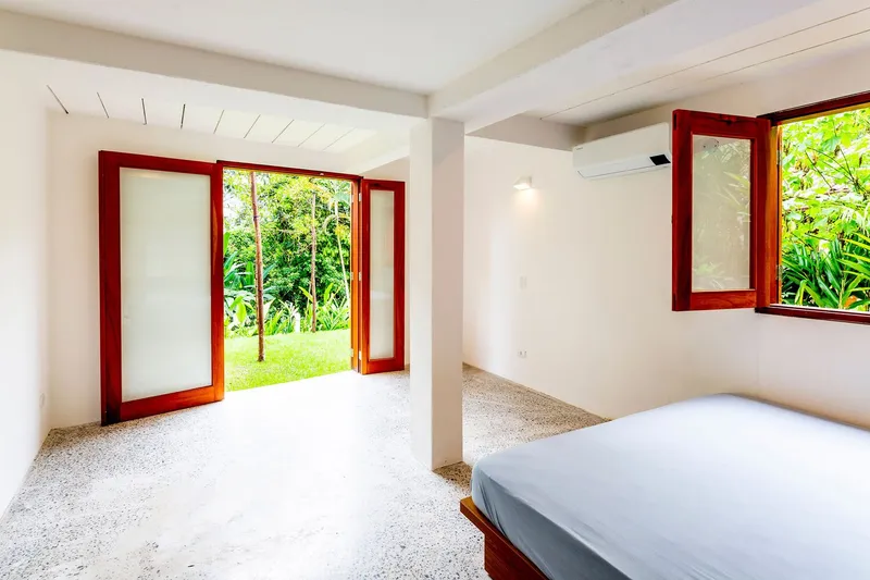 Bedroom interior with red timber framed doors and windows opening directly onto a lawn with tropical plantings