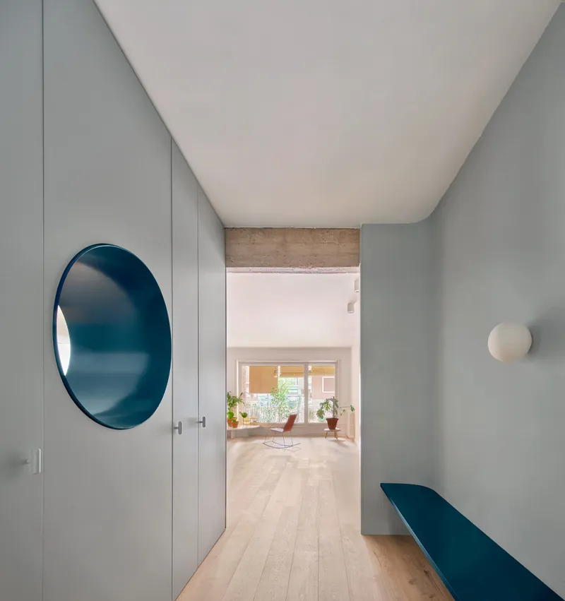 Hallway with pale wood flooring, grey walls, a circular blue mirror, and view through to sunlit living space