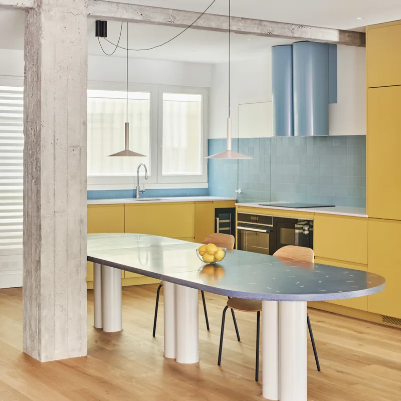 Kitchen with yellow cabinetry, light blue tile backsplash, exposed concrete beam, and oval dining table on white cylindrical legs