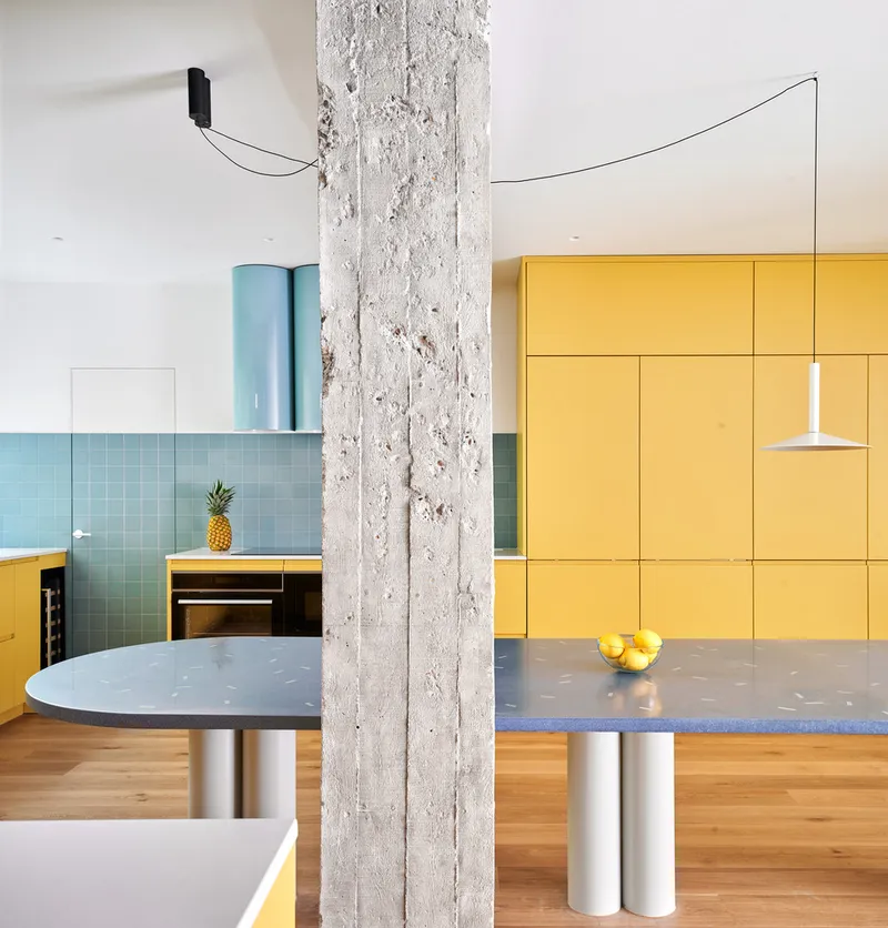View across kitchen showing exposed concrete column, yellow storage wall, blue terrazzo countertop, and suspended pendant light