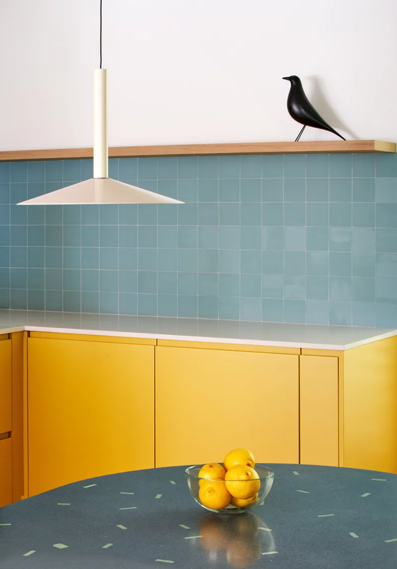 Detail of kitchen backsplash with small-format blue tiles, yellow lower cabinets, and pendant fixture above terrazzo countertop