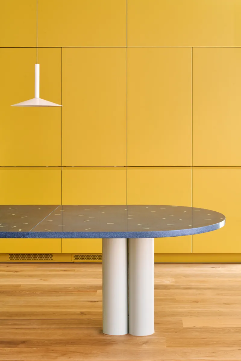 Round terrazzo table with twin cylindrical white legs against a yellow tile wall with light oak flooring