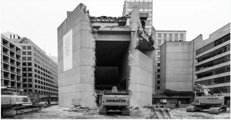 Partially demolished concrete structure with exposed interior and excavators positioned at its base surrounded by office buildings