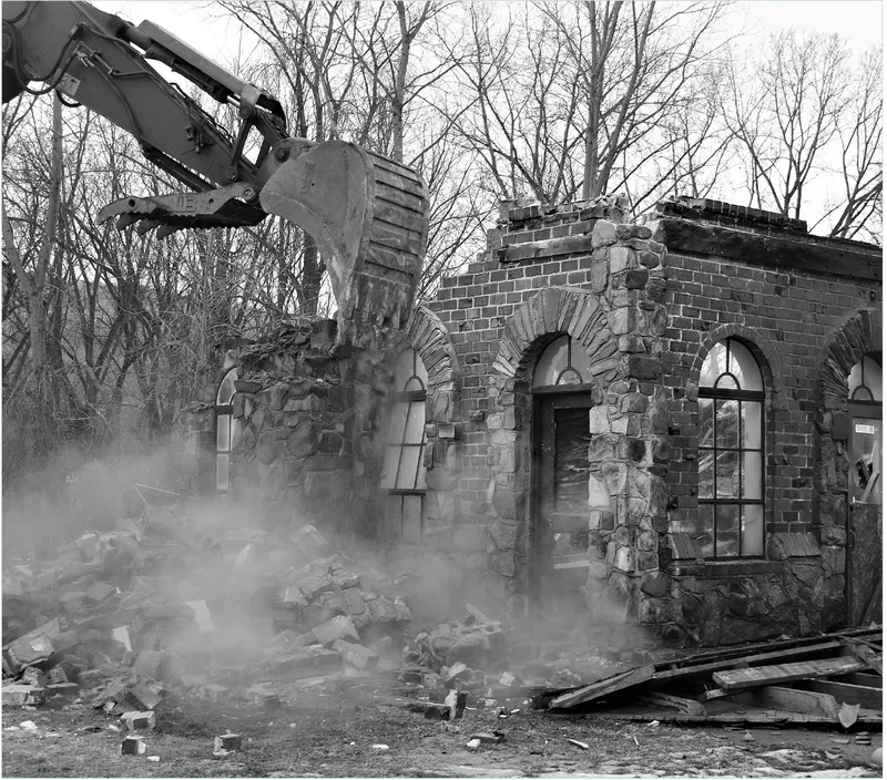 Excavator demolishing a brick building with arched windows amid dust and bare winter trees