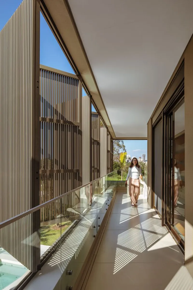 Glass-enclosed walkway with vertical timber slat screen casting shadows as a woman walks through in sunlight