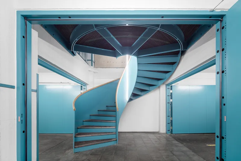 Turquoise spiral staircase rising through a white atrium with exposed timber ceiling above