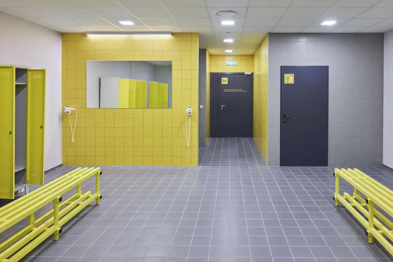 Locker room interior with yellow tiled wall, grey floor tiles, yellow benches, and dark doors