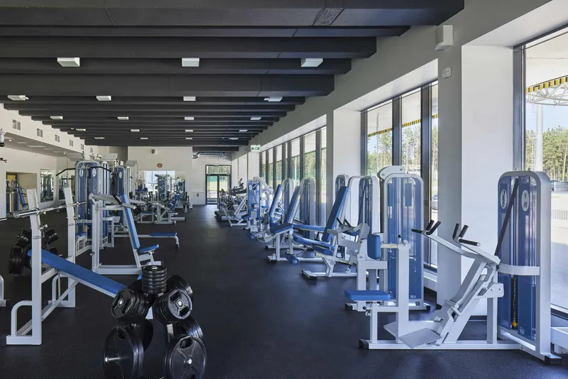Interior fitness facility with rows of exercise equipment beneath exposed black ceiling beams and floor-to-ceiling windows