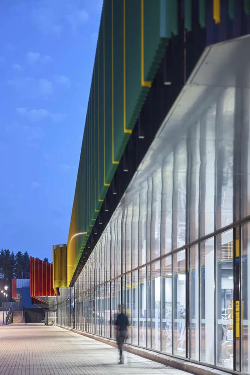 Exterior walkway along vertical corrugated metal cladding and floor-to-ceiling glazing at dusk with a figure passing