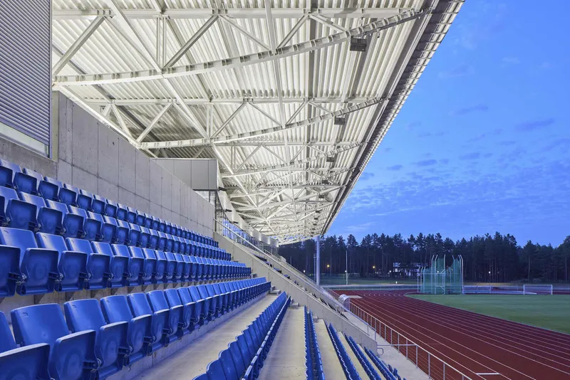 Stadium seating with blue plastic seats facing an athletics track under a white steel truss canopy at dusk