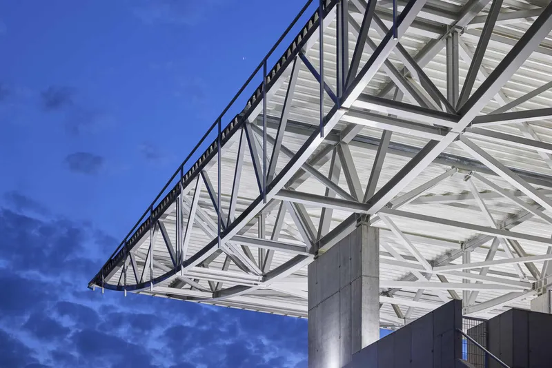 Underside of the cantilevered white steel truss roof supported by concrete columns against a twilight sky