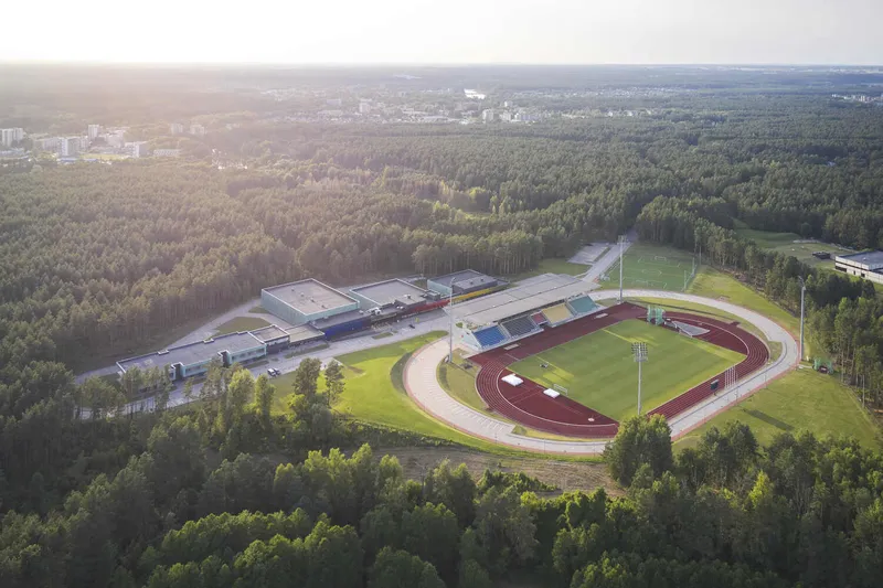 Aerial view of an athletics stadium and track surrounded by forest with a distant urban edge