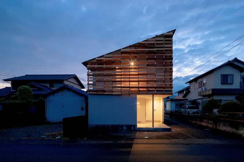 Street view of the horizontal timber screen facade illuminated at dusk between neighboring houses