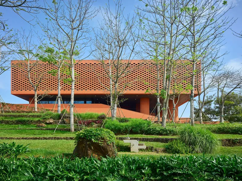 Perforated terracotta facade elevated above terraced landscaping with young deciduous trees in spring