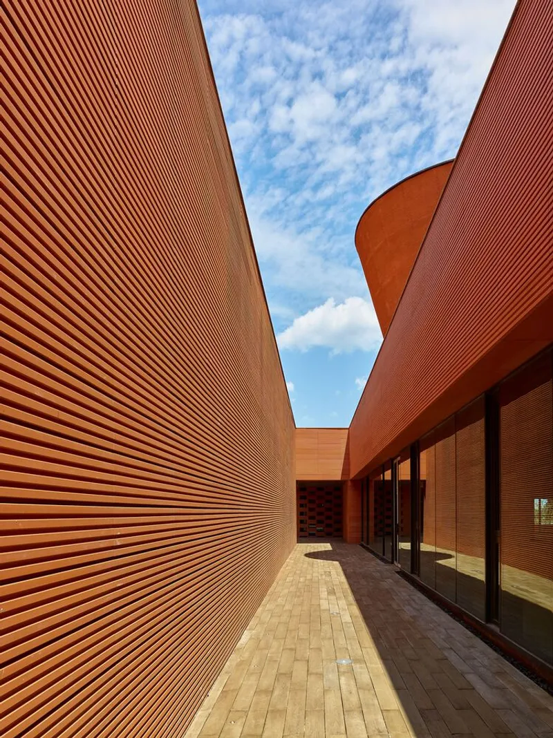 Narrow courtyard between ribbed terracotta walls and glazed facade under blue sky with clouds