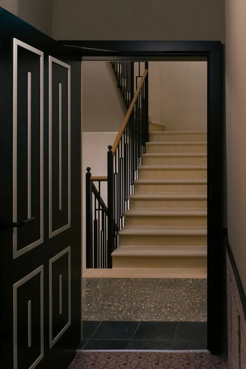 View through a black paneled door to a timber staircase with vertical metal railings and terrazzo flooring