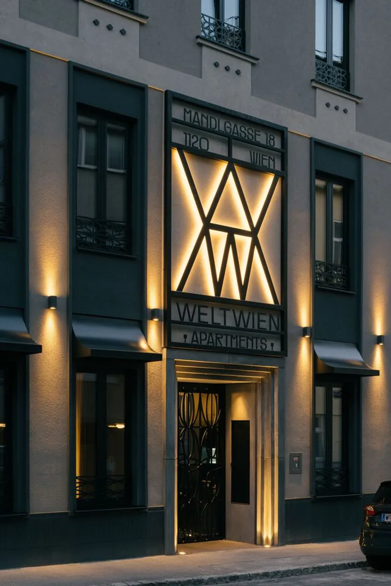 Street facade with geometric illuminated panel above the entrance and vertical wall sconces at dusk