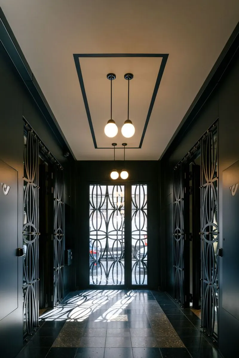 Corridor with dark paneled walls and patterned metal screens on entry doors filtering daylight onto the floor