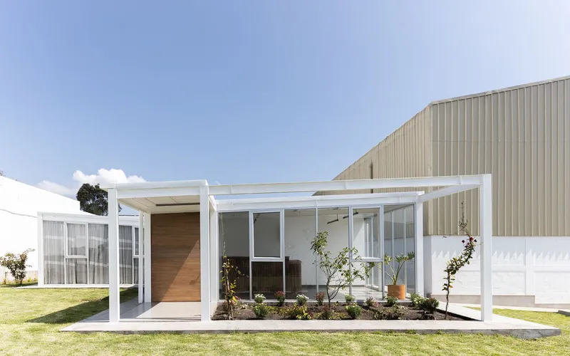 White steel frame pergola with timber entry door and full-height glazing overlooking planted garden bed