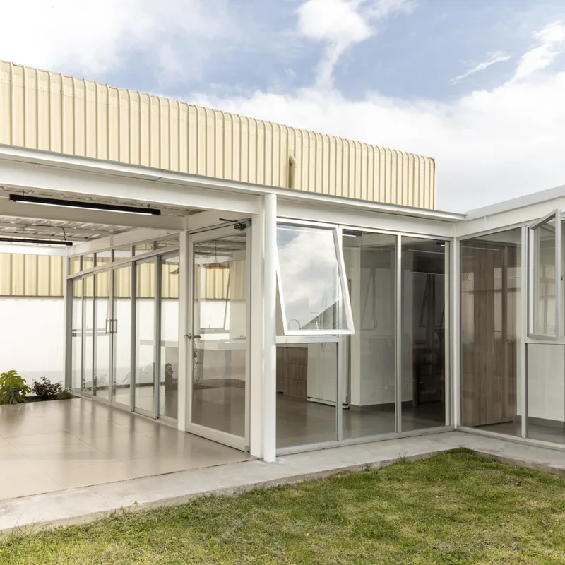 Covered walkway with white steel frame and glass sliding doors connecting volumes under corrugated metal cladding