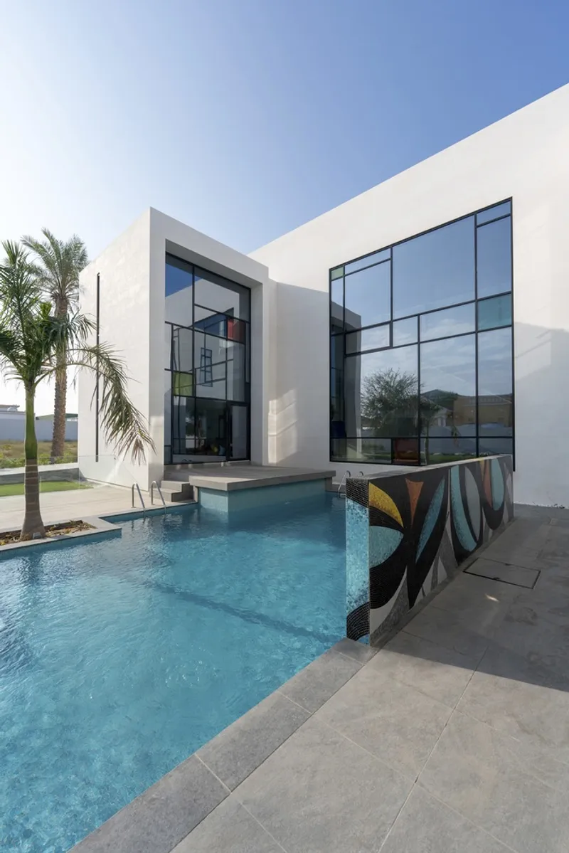 White stucco facade with floor-to-ceiling glass walls overlooking a blue pool with palm trees and grey paving