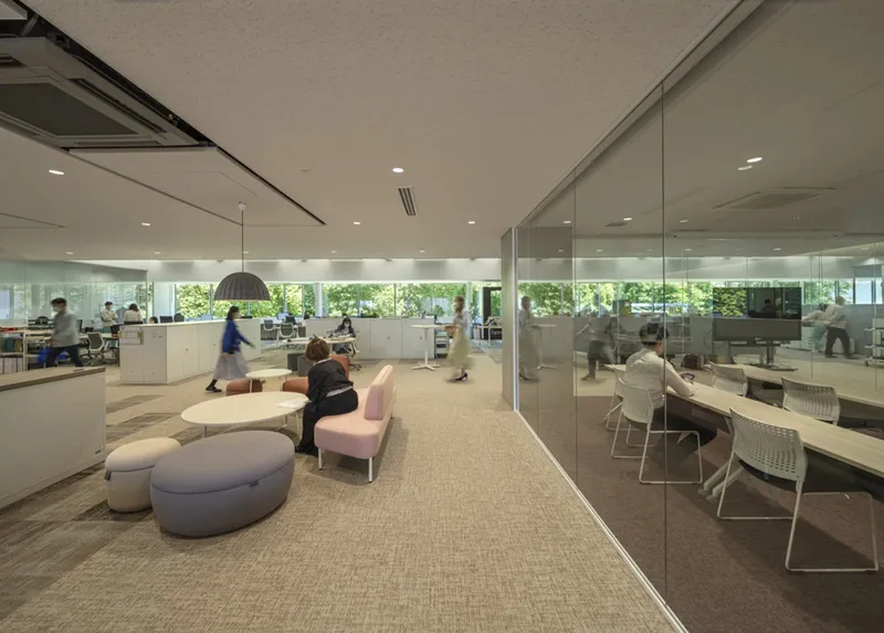 Lounge seating zone with upholstered ottomans adjacent to a glass-walled meeting room and horizontal ribbon windows framing greenery