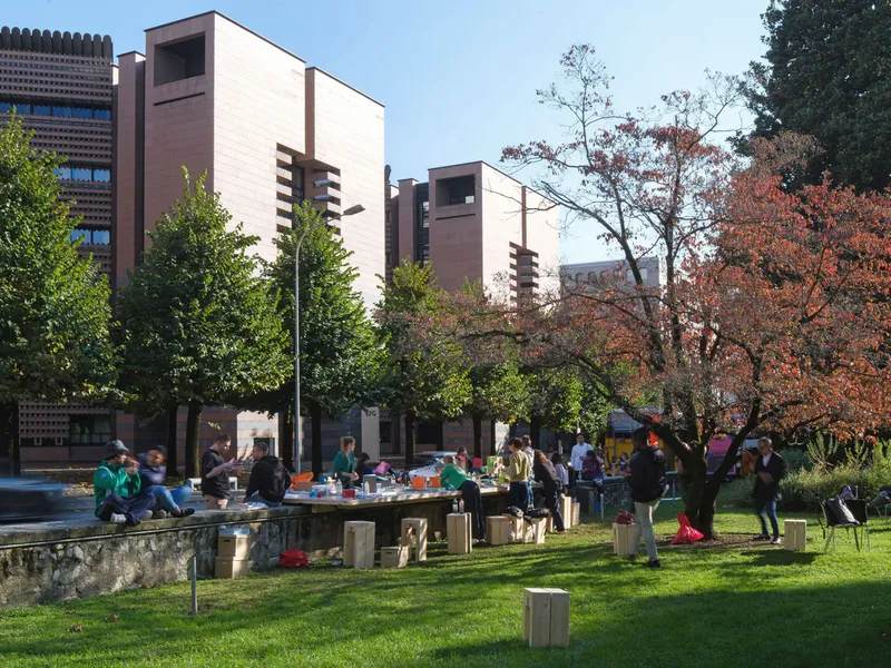 People gathered around a long timber table in a park with pink masonry buildings behind