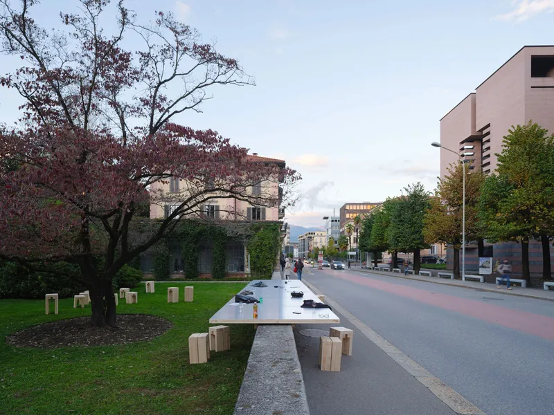 Stainless steel communal table with timber stools along a pedestrian boulevard under autumn trees at dusk