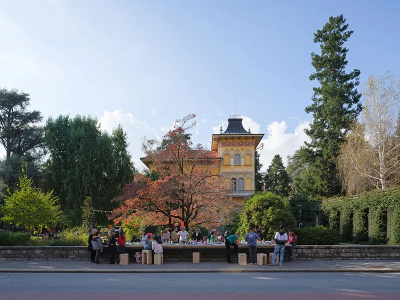 Public gathering around the long metal table installation with a yellow villa tower visible beyond the trees