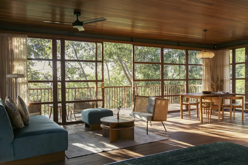 Open living and dining space with timber ceiling and gridded glass doors opening to a balcony amid trees