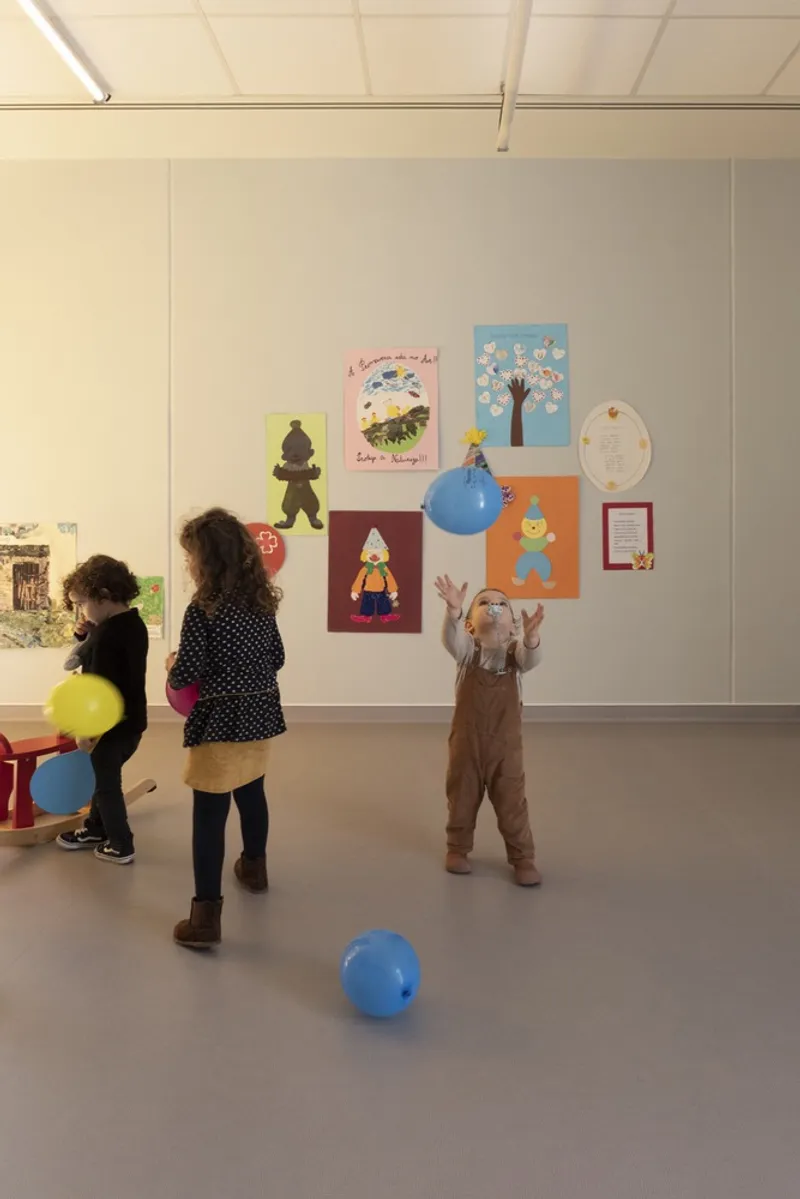 Interior classroom with white walls displaying children's artwork and three children playing with blue balloons