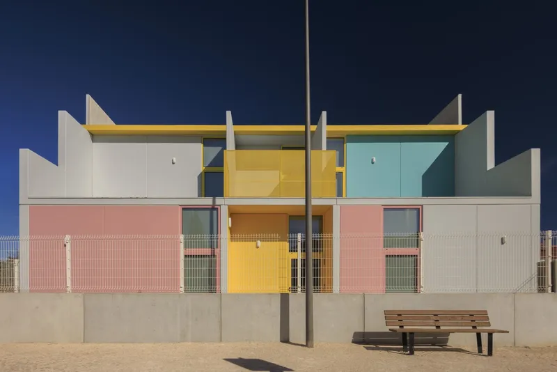 Symmetrical facade with yellow and turquoise upper panels above pink roller shutters and a wooden bench