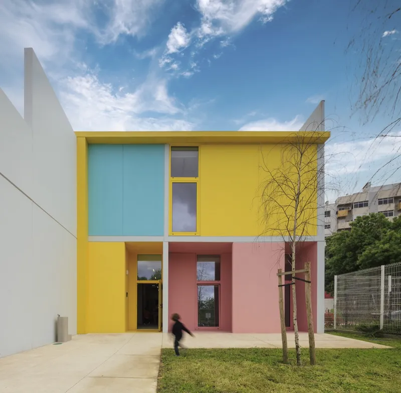 Front elevation showing yellow and pink panels with young trees in the foreground under clouds