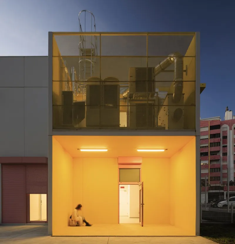 Two-story glass-fronted volume with yellow interior illumination and exposed industrial ductwork at dusk