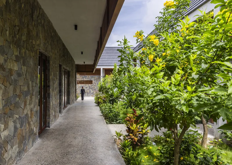 Outdoor corridor along a stone wall with dense tropical plantings and a figure walking ahead