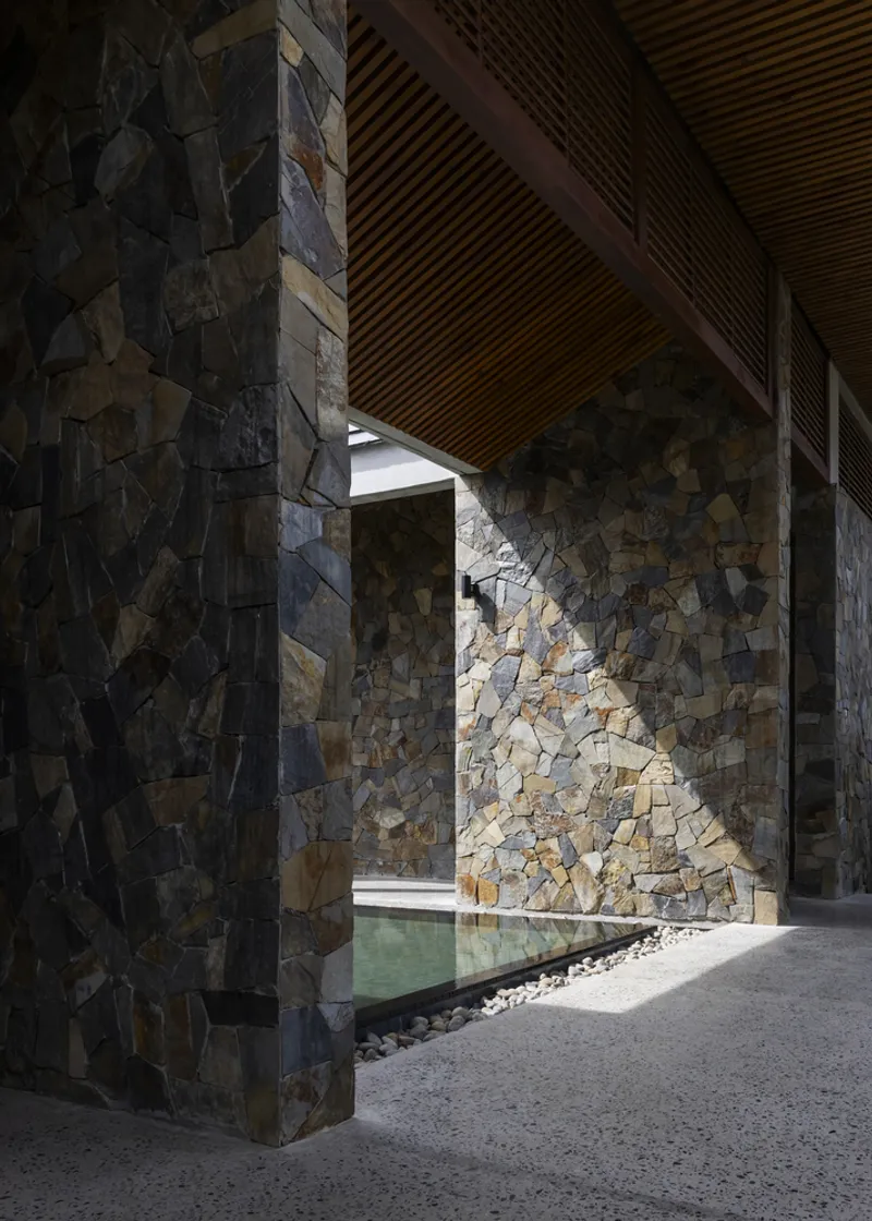 Entry passage framed by irregular stone walls with timber slatted ceiling and reflecting pool beyond