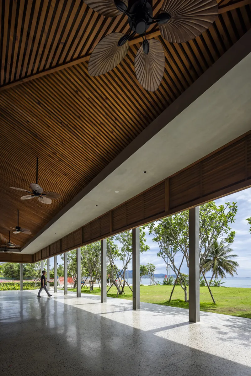 Open pavilion with timber slatted ceiling and terrazzo floor overlooking a lawn with coastal palms and a figure walking