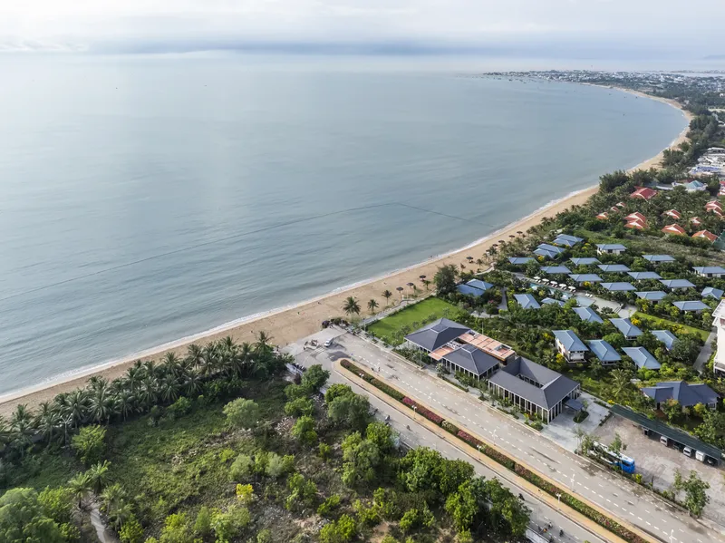 Aerial view of low-rise buildings with gabled roofs nestled between palm trees and a sandy beach