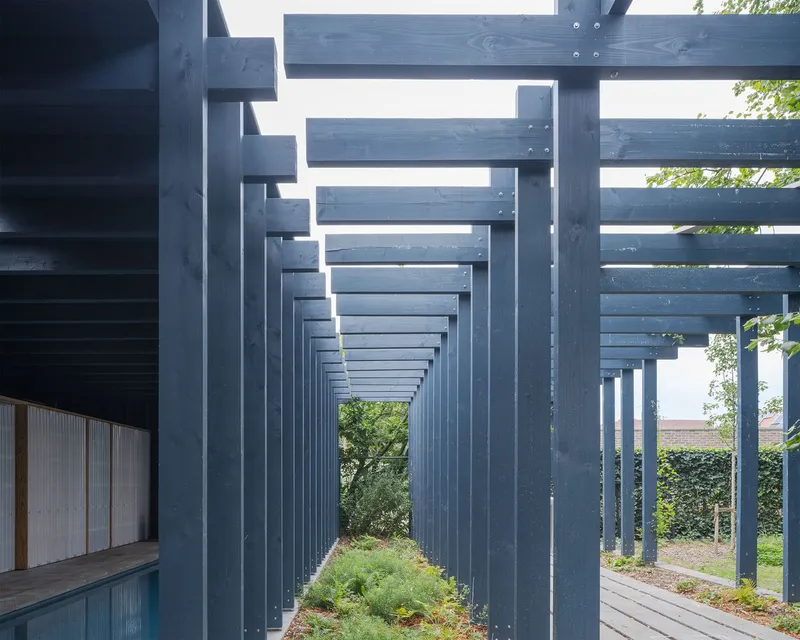 Linear timber colonnade with repeated blue-grey posts creating a rhythmic passage alongside vegetation