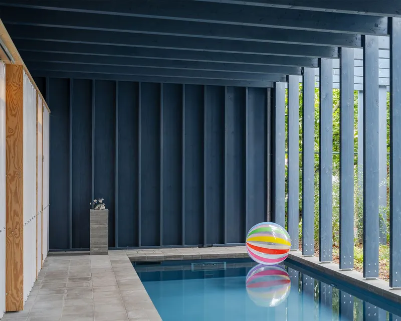 Covered poolside terrace with vertical dark metal fins filtering sunlight and green foliage beyond