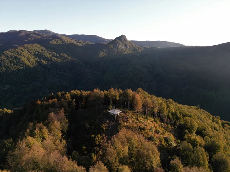 Aerial view of a cruciform pavilion nestled atop a forested hilltop with autumn foliage and distant peaks