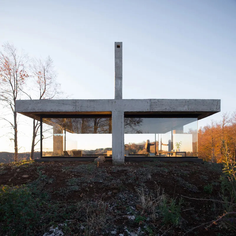 Elevated concrete pavilion with cruciform plan and central chimney on a cleared earthen plateau in autumn