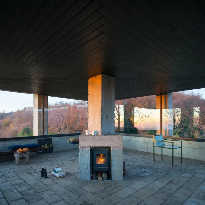 Interior terrace with central concrete fireplace beneath a dark timber plank ceiling at dusk